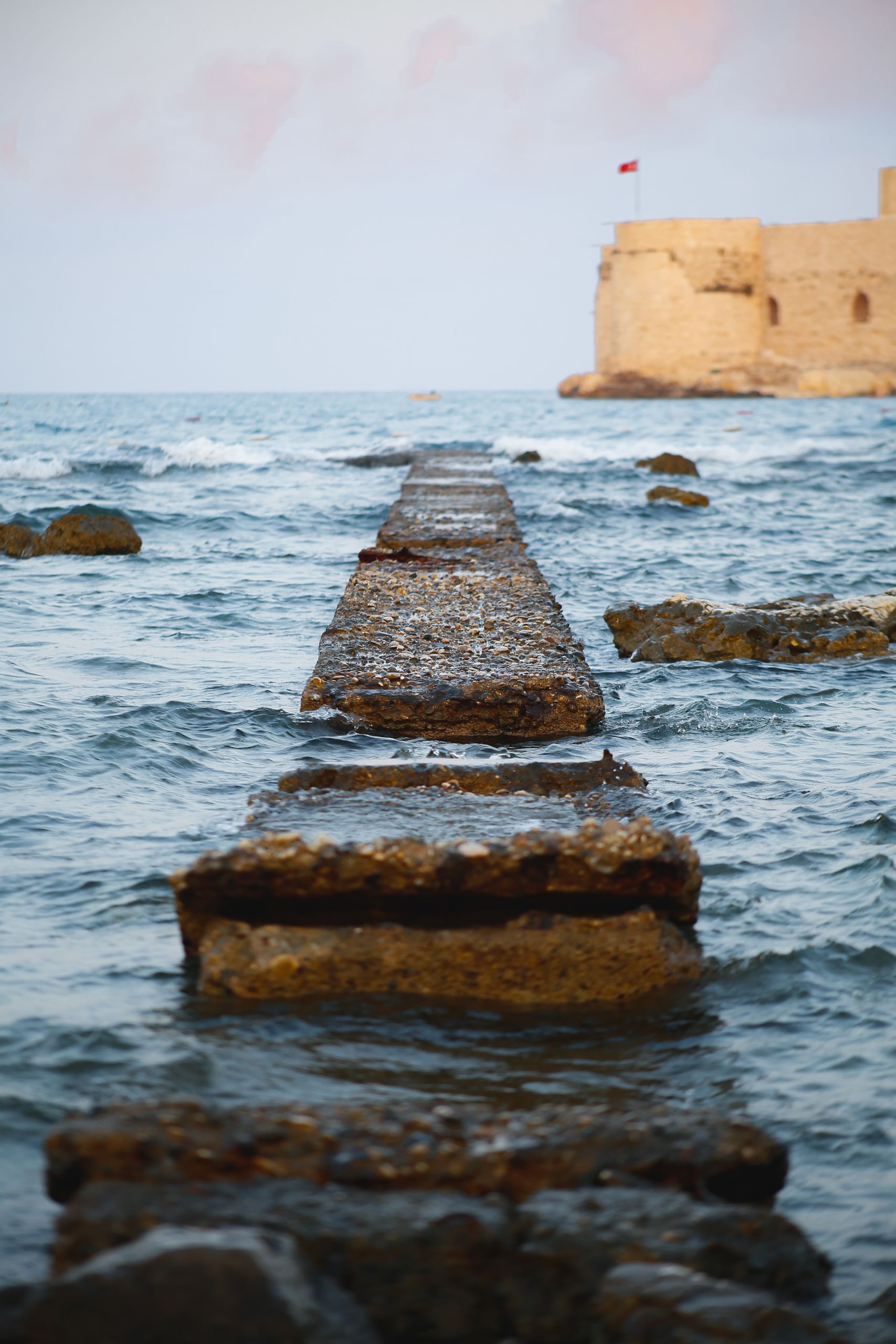 A stone jetty stretches out to a historic seaside castle under the bright sky.